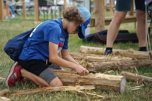A child shaves the bark off a log with a tool.