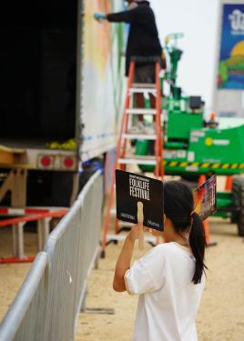 A young girl stands with two paper fans near her head, one reading “Smithsonian Folklife Festival.” She stands near a fence, looking into a large trailer awaiting a mountain biking demonstration.