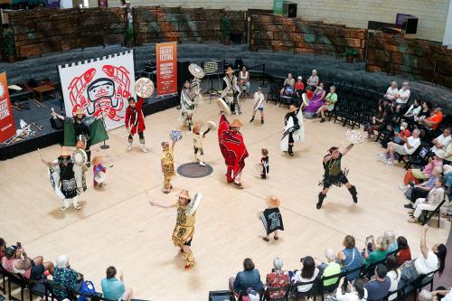 A group wearing Tsimshian regalia dances in the center of a circular performance space, with audiences circling around them. All performers, except the youngest children, raise up circular hand drums and beaters.