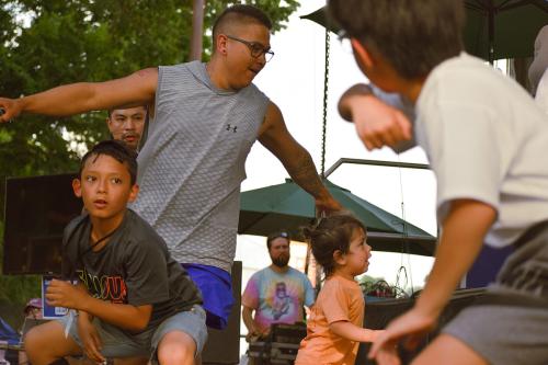 Young children, a toddler, and young men dance together in front of the stage. Some are crouched, and one has his arms outstretched like a bird. 