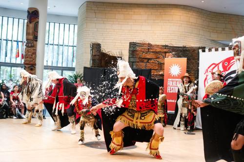 The dance group performs indoors, lined up wearing matching black and red capes, leather boots, and white headdresses. A plume of white feathers flies through the air.