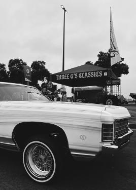 A white lowrider parked outdoors, next to a tent that reads Three G's Classics. Black-and-white photo.
