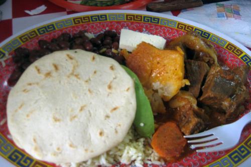 Rice, vegetables, meat, and a pupusa on a plate with a Chinese pattern around the rim and a plastic fork sticking out.