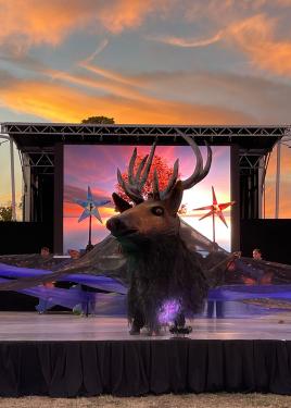 The elk head puppet, again, at center stage, under bright pink clouds in a dark blue sky. A screen at the back of the stage seems to show the same sky.