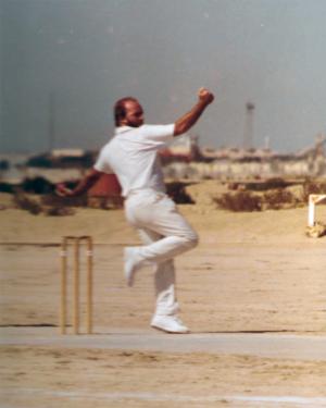 A man in a dirt field prepares to pitch a ball. Behind him is a blurred horizon of shrubbery and buildings. 