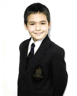 A young boy wearing a black blazer with school logo and tie smiles slightly, standing against a white backdrop..
