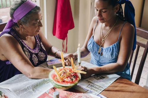 Two women, one an elder, close their eyes and embrace hands over a kitchen table. Between them on the table is a lit candle and half a red watermelon with various types of flowers stuck into its flesh.