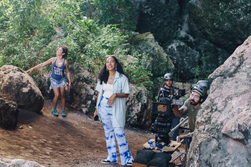 Two women stand outdoors on a dirt path flanked by boulders. Three others stand to the side, all in black helmets, operating a high-end film camera.