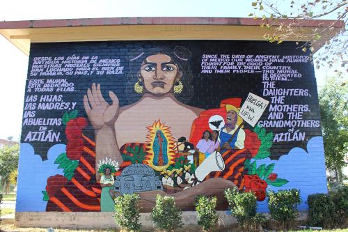 Mural on the side of a brick building, featuring a looming brown-skinned figure with gold earrings and one open hand raised. In their other arm, the person holds smaller figures, including Guadalupe, farmworkers with a Huelga! VIVA UFW sign raised, worker