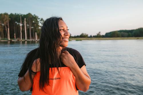 A Black woman wearing black T-shirt and orange waders smiles, looking off into the distance, standing in front of a blue body of water with green trees in the distance. 
