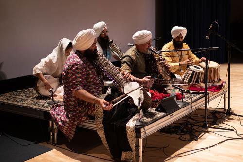 Five Sikh men and one Sikh woman sit on stage. They sit cross-legged on a raised platform. They play various stringed instruments and a drum.