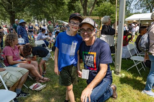 A man poses and smiles with a young boy amid a crowd of people sitting and standing in a grass lawn; the man wears a grey baseball cap and dark blue T-shirt with 2025 Smithsonian Folklife Festival's theme logo, Youth and the Future of Culture; the boy wea