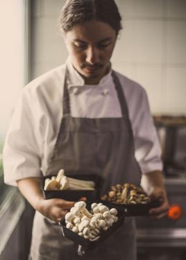 Man in white chef's coat and gray apron looks down at the three containers of mushrooms he holds.