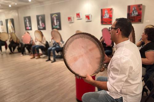 Around the perimeter of a room, people sit playing large, round frame drums.