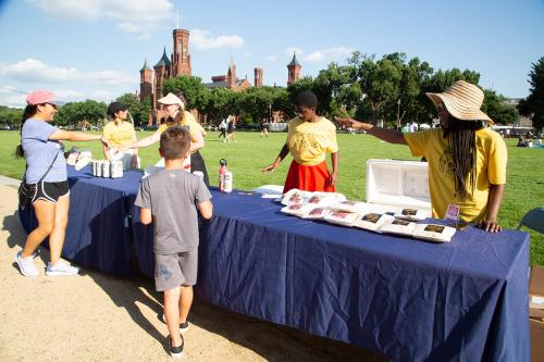 Four individuals in matching yellow shirts pass out drinks and bags of popcorn to two visiting individuals at an outdoor table. The table is covered with a blue cloth and behind the table is a grass lawn, trees, and a large brick building. 