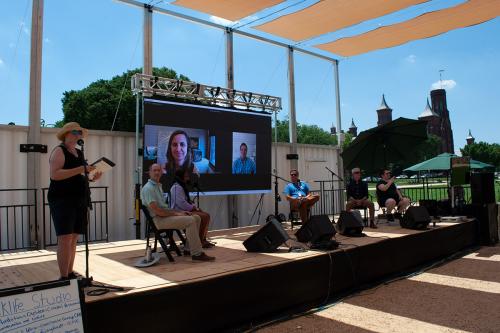 Six people seated on an outdoor stage, including an ASL interpreter, plus two people on screen via Zoom.