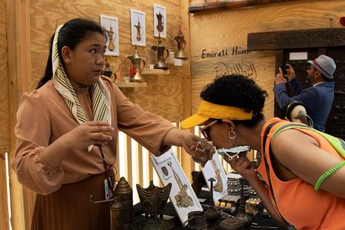 A woman holds up a small glass with grounded spice in it to another woman who is leaning over to smell. Behind them, a man takes a picture of golden pots hanging on the wall near words  “Emirati Home.”