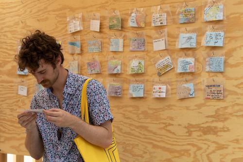 A man inspects a piece of paper in his hands, standing in front of a plywood wall containing other small pieces of paper: bits of colorful maps with handwritten notes on them.