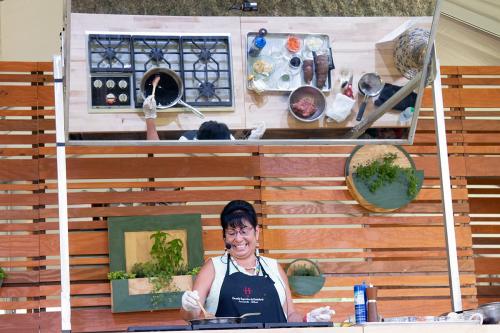 A woman smiles and stands over a stove, stirring a sauce in a pan that is reflected in a mirror above her. Also reflected in the mirror are other ingredients and cooking utensils.