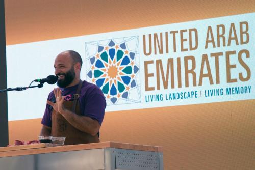 A man in a leather apron laughs and holds up one hand, fingers splayed, talking into a microphone on stage. Behind him, a digital backdrop with the logo: United Arab Emirates, Living Landscape, Living Memory.