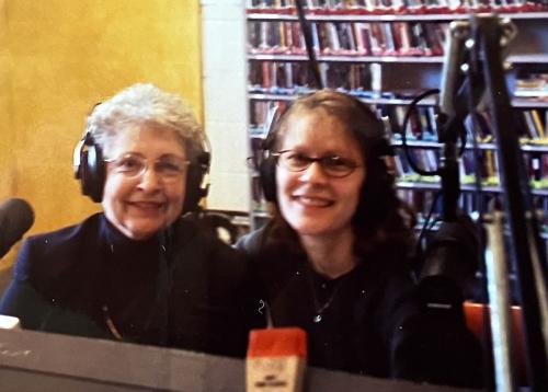 An older and younger woman sit at a radio control booth with headphones and microphones, smiling from behind the glass.