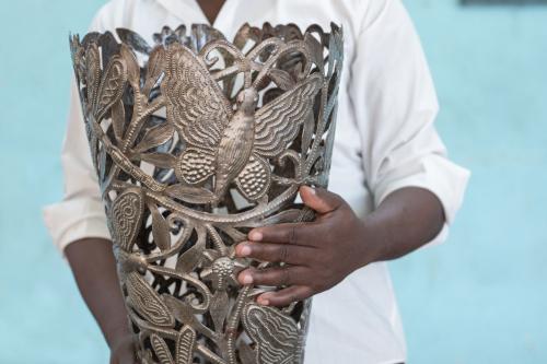A Black man in a white, button-up shirt holds a tall, decorative metal vase. Cut-outs in the vase reveal a design of butterflies and flowers.
