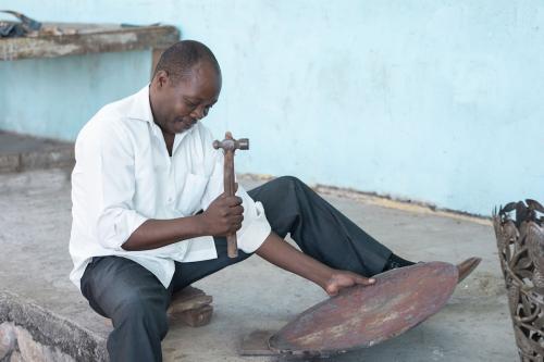 A seated Black man holds a sheet of metal shaped like a disc in one hand and a mallet in the other. Halfway out of frame to the right of the man is a decorative metal vase.
