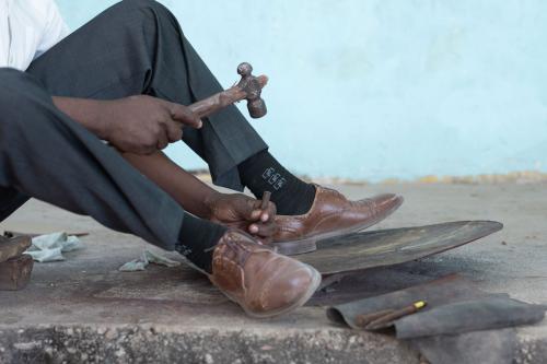 A seated Black man whose upper body is out of frame chisels at a sheet of metal shaped like a disc with a mallet.