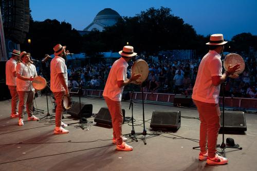 Five men in matching white shirts, khaki pants, white sneakers, and tan hats plays hand drums and sing on stage for a crowd of people.