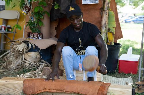 A man sits on the ground, hitting a rectangular piece of bark with a wooden mallet.
