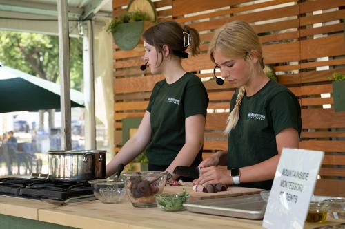 Two young women in matching forset green Mountainside Montessori T-shirts stand at an outdoor demo kitchen. One is cutting root vegetables with a knife.