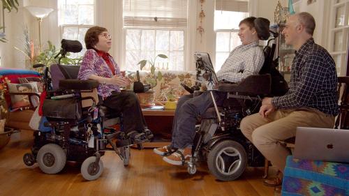 Film still of an elder woman and a young man, both in wheelchairs, facing each other in conversation and smiling in a warmyl lit living room. A man sits to the side, next to a laptop, also smiling.