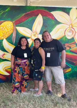 Two women and a man pose in front of the vanilla blossoms of the mural. One woman wears a black apron that is splattered with paint.