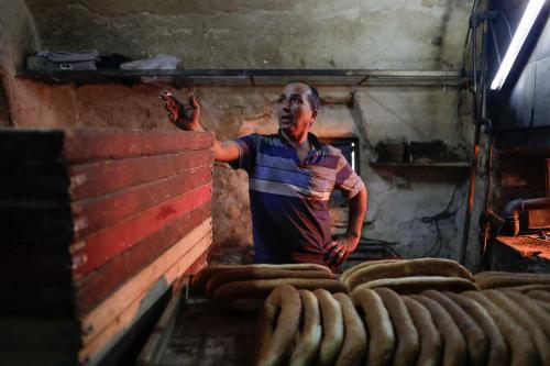 A man stands behind a tray of freshly baked ring-shaped bread. He is lit by a glowing red oven just out of from to the right.