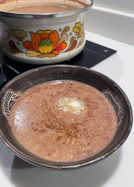 A bowl of thin brown porridge, enxt to a vintage pot filled with more of the porridge.