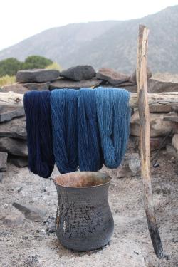 Three shades of blue-dyed yarn hanging over a wooden rack above a metal pot, with mountains in the background.