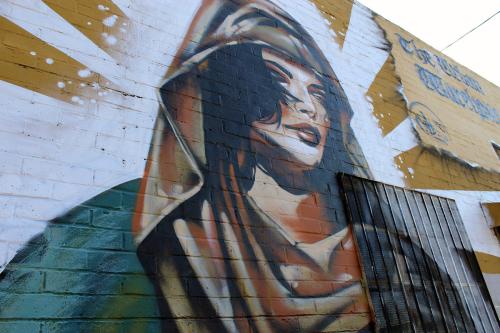 Painting on the exterior of a brick building, with the image of woman with dark hair, teal shawl and reddish head covering. Her image overlaps a window grate.