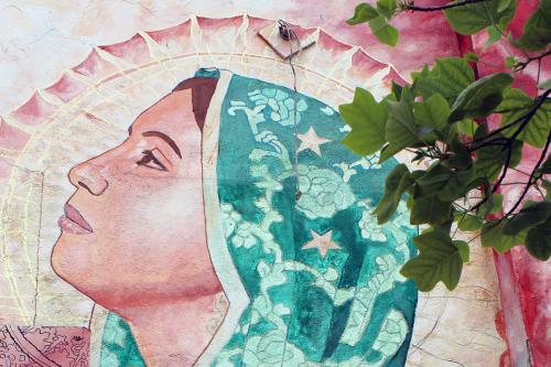 Outdoor painting, partly obscured by green leaves of a tree. It features the virgin figure in pink and teal.