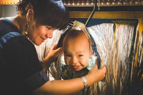 A baby sticks his head through the vertical warp of yarn on a loom, held open by a young women who smiles at him.