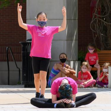 Two dancers in matching pink shirts performing on asphalt outdoors, one seated and hunched over, the other standing with arms raised.