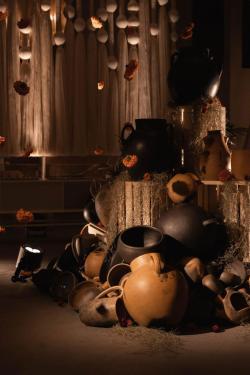 Artistic display of ceramic jugs and vases in various brown tones, arranged in stacks among hanging ornaments and dramatic lighting.