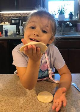 A young girl holds up a small round of dough, leaning on a kitchen counter.