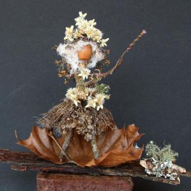 A human-like figure made of white and yellow flowers for a crown, white fuzz for hair, an acorn for a head, and twigs fora body and flared skirt. It stands on a brown maple leaf and waves a wooden wand.