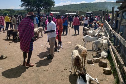 A row of brown and cream-colored goats are tied to a wooden fence, while people in modern and traditional Kenyan dress, like bright red and blue woven shawls, talk and shop.