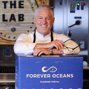 Headshot of a man with white hair and beard, smiling as he rests his arms on a blue cardboard box that reads Forever Oceans
