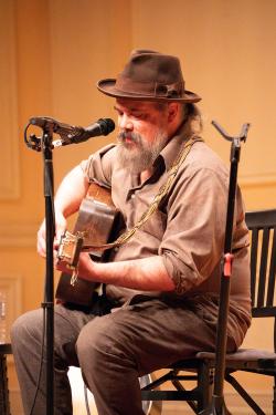A man in a brown fedora and gray dress shirt, with a gray beard, plays acoustic guitar on stage.