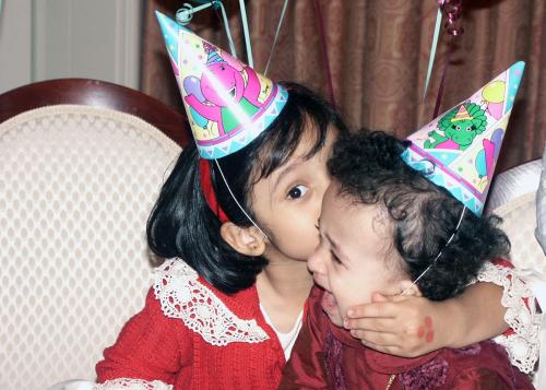 A young girl stares at the camera with one eye obscured by her younger sister's head as she whispers into the sister’s ear. The two girls wear red.