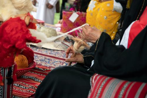 Two hands quickly spin white wool into yarn in the foreground. The woman spinning is seated on colorful carpet next to red and yellow yarn in a basket. 