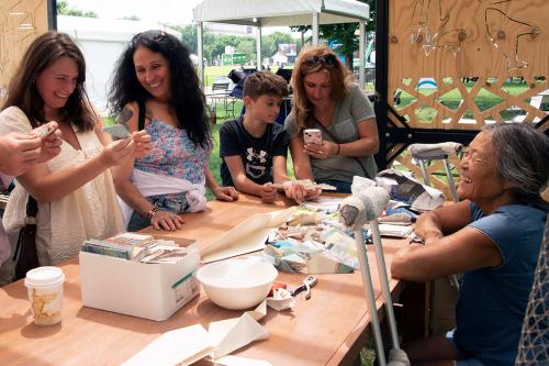 A seated woman with crutches beside her smiles at four people standing around a wood table in an outdoor wooden construction. The table is littered with folded paper and the standing individuals are folding origami.