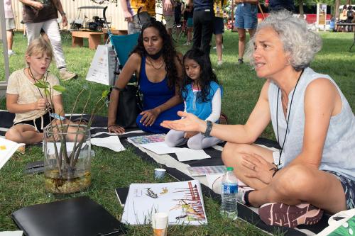 Two small kids and two adults sit in on mats on the ground with watercolor paints and paper. The woman in the foreground, with gray hair and gray top, gesticulates as she explains something. In the center of their circle, a glass vase holding mangrove pla